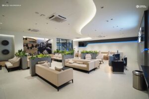 Modern showroom lounge with cream sofas, green plants, and car-themed decor; bright lighting and sleek, minimalist design.