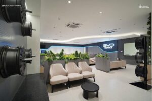 Modern car service lounge with beige chairs, small black table, potted plants, wall-mounted tire rims, and BYD Service signage on the back wall.