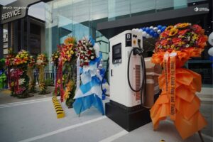 A white electric vehicle charging station stands in front of a building entrance, surrounded by colorful flower arrangements and ribbons.