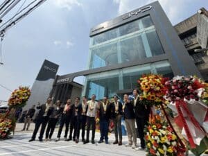 A group of people pose in front of a BYD car dealership building with large flower stands on both sides under a bright, sunny sky.