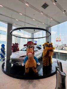A white car is displayed on a circular platform indoors, with people in animal and cowboy costumes standing in front. Balloons and large windows are visible in the background.