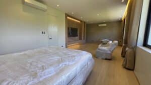 Spacious bedroom with a bed, sofa, and chair all covered in protective plastic, a wall-mounted TV, wooden floors, large window, and recessed lighting.