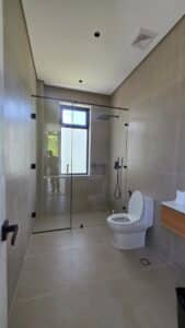 Modern bathroom with beige tiles, glass-enclosed shower, white toilet, wall-mounted sink, and a window letting in natural light. A person is visible in the mirror’s reflection.
