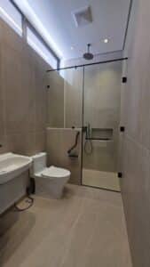 Modern bathroom with beige tiles, wall-mounted sink, toilet, and a glass-enclosed shower featuring a rain shower head, grab bars, and a handheld shower.