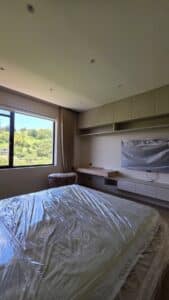 A modern bedroom with a large window, built-in desk, and shelves. Furniture, including the bed and chair, is covered in protective plastic. Greenery is visible outside the window.