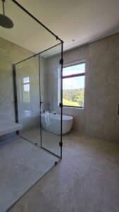 Modern bathroom with beige tiles, a glass-enclosed shower, a freestanding bathtub by the window, and natural light entering through the window.