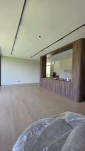 A spacious, modern room with light wood flooring, a large wooden partition with a countertop, minimal furnishings, and large windows letting in natural light.