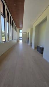 A modern, empty room with light wood flooring, tall windows along one wall, and some stacked tiles in the corner.