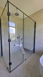 A modern glass shower enclosure with black frames, a wall-mounted shower head, and a control handle covered in protective plastic. A window outside is visible in the reflection.