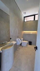 Modern bathroom with beige tiles, a pedestal sink, wall-mounted urinal, toilet, black trash bin, large mirror, recessed shelf with lighting, and a high window.
