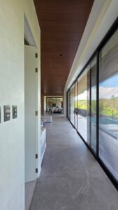 A modern hallway with large glass windows on the right, a wooden ceiling, gray stone floor, and a distant view of a dining area.