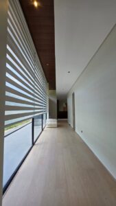 A modern hallway with light wood flooring, large floor-to-ceiling windows with striped blinds, and a white armchair at the end.