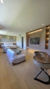 A spacious, modern living room with furniture covered in plastic, wooden floors, built-in shelves, and a large television also covered in plastic.