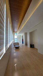 A modern, minimally furnished room with wood flooring, large windows with blinds, two armchairs, and recessed ceiling lighting.