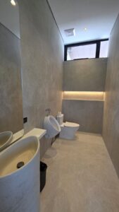 Modern restroom with a sink, urinal, toilet, and recessed wall lighting. The space features large grey tiles and a mirror on the left wall.