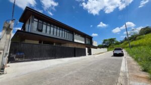 A modern two-story building with large windows and a black gate is situated along a sloped, paved street under a bright blue sky with scattered clouds.