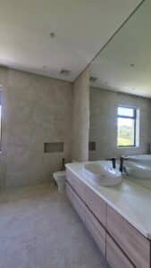 Modern bathroom with beige tiles, a wall-mounted toilet, a floating vanity with a sink, and a large mirror reflecting a window with a view outside.
