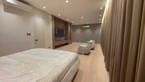 A modern bedroom with light wood floors, a bed and chairs covered in plastic, built-in shelves, and large curtains along one wall.