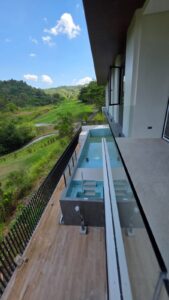 View from a balcony overlooking a small private pool, wooden deck, and fence, with green hills and a clear blue sky in the background.