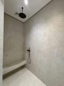 Minimalist shower with beige tiles, a built-in bench, a black ceiling-mounted rainfall showerhead, and a handheld showerhead on the wall.