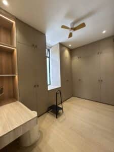 Modern, well-lit room with beige built-in wardrobes, a wood desk with shelves, a window seat, step stool, and ceiling fan. The floor is light wood, and the lighting is recessed.