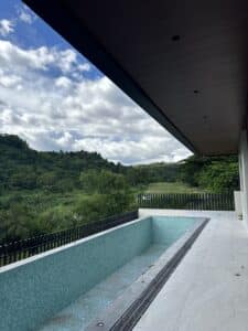 An empty rectangular swimming pool on a tiled patio overlooks green hills and trees under a partly cloudy sky.