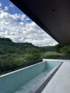 Empty outdoor pool with turquoise tile next to a patio, overlooking green hills and trees under a partly cloudy sky.