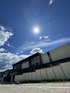 A modern building with large windows under a bright sun and blue sky with scattered clouds; some items are on the ground near the wall.