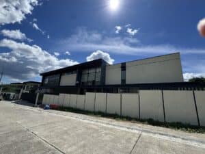 A modern two-story building with large windows and a flat roof is pictured under a bright blue sky with sun and clouds, behind a tall pale fence along a concrete road.