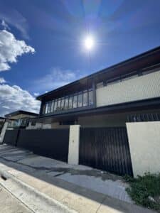Two-story modern house with large windows and a black metal gate, photographed from the sidewalk on a sunny day with a bright sun and blue sky.