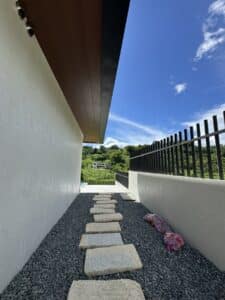 A narrow stone pathway runs alongside a white building, bordered by gravel and a black metal fence, with greenery and blue sky in the background.