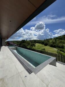 Long rectangular swimming pool on a tiled patio, overlooking a green landscape and partly cloudy sky, with a black fence along the edge.