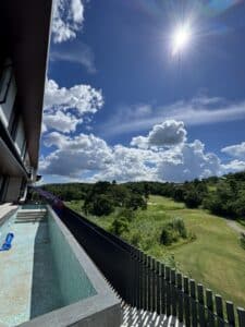 View from a balcony with a pool overlooking a green landscape and trees under a bright blue sky with scattered clouds and the sun shining.