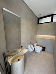Modern bathroom with a vessel sink, wall-mounted faucet, large mirror, urinal, toilet, and a black waste bin. Beige tiles cover the floor and walls, with overhead and accent lighting.