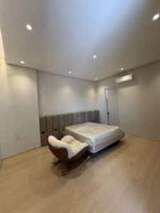A minimalist bedroom with a mattress on a bed frame, a cushioned chair, wood flooring, white walls, and ceiling lights. An air conditioner is mounted above a closed door.
