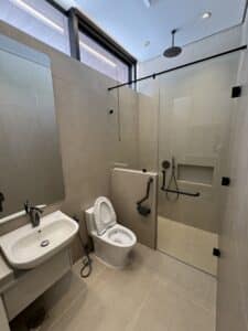 Modern accessible bathroom with a wall-mounted sink, toilet with grab bars, and a glass-enclosed shower featuring a rainfall showerhead and handheld sprayer.