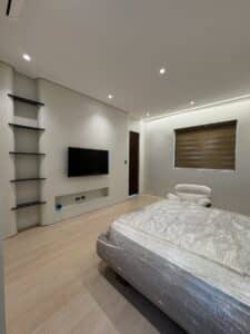 Minimalist bedroom with light wood flooring, a bed wrapped in plastic, wall-mounted TV, black shelves, a white chair, and a window with a brown blind.