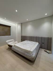 Minimalist bedroom with an unmade bed covered in plastic, a lounge chair, padded wall paneling, wooden blinds, and light wood flooring.