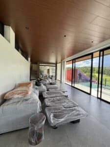 Modern living room with large windows, unused furniture covered in plastic wrap, and a view of green landscape and a red tent outside.