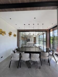 Modern dining room with a glass table, eight chairs wrapped in plastic, pendant lights above, golden wall decor, and large windows offering views of greenery.