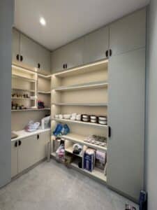 A modern pantry with light cabinets, open shelves holding dishes, food items, and kitchen supplies, under built-in ceiling lights.