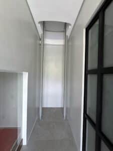 A narrow hallway with gray tile flooring and white walls, ending in a vertical lift or dumbwaiter shaft with visible metal rails.