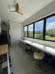 A bright kitchen with large windows, two bar stools at a counter, a ceiling fan, and a view of green fields and blue sky outside.