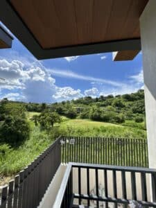 View from a balcony showing a wooden fence, green grassy landscape, trees, and a blue sky with scattered clouds.
