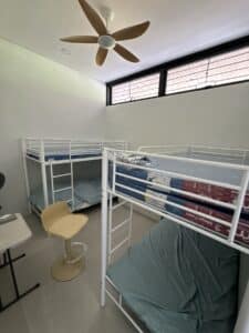 A small room with two white metal bunk beds, each with blue sheets, a yellow chair, a desk, and a ceiling fan with wooden blades.