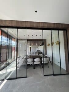 Modern dining room with a long table and chairs covered in plastic, viewed through large glass sliding doors; kitchen area visible in the background.