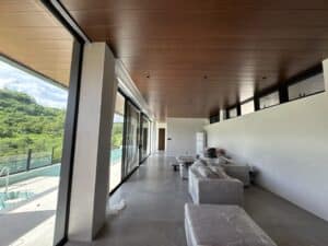 Modern living room with large windows, wood-paneled ceiling, and furniture covered in plastic; view of pool and greenery outside.