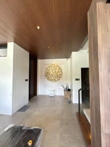 A modern interior with beige tile flooring, wood-paneled ceiling, and a gold circular wall sculpture. A step ladder and covered furniture indicate ongoing setup or renovation.