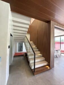 Modern indoor staircase with white steps, a black handrail, wooden accents, and recessed step lighting, located in a bright room with large windows.