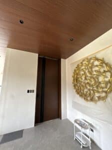 Modern entryway with a tall wooden door, tiled floor, white walls, abstract gold wall art, light switches, and a small white cart.